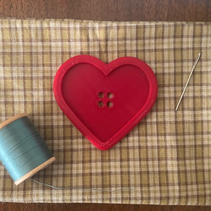 Heart-shaped button, spool of thread, and needle on a checkered cloth with a blue plate in the background.