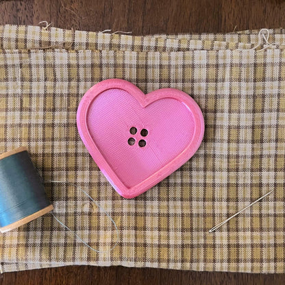Heart-shaped pink button on a checkered fabric with a spool of blue thread on a wooden surface.