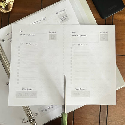 Person cutting a printed planner sheet with green scissors on a wooden surface.