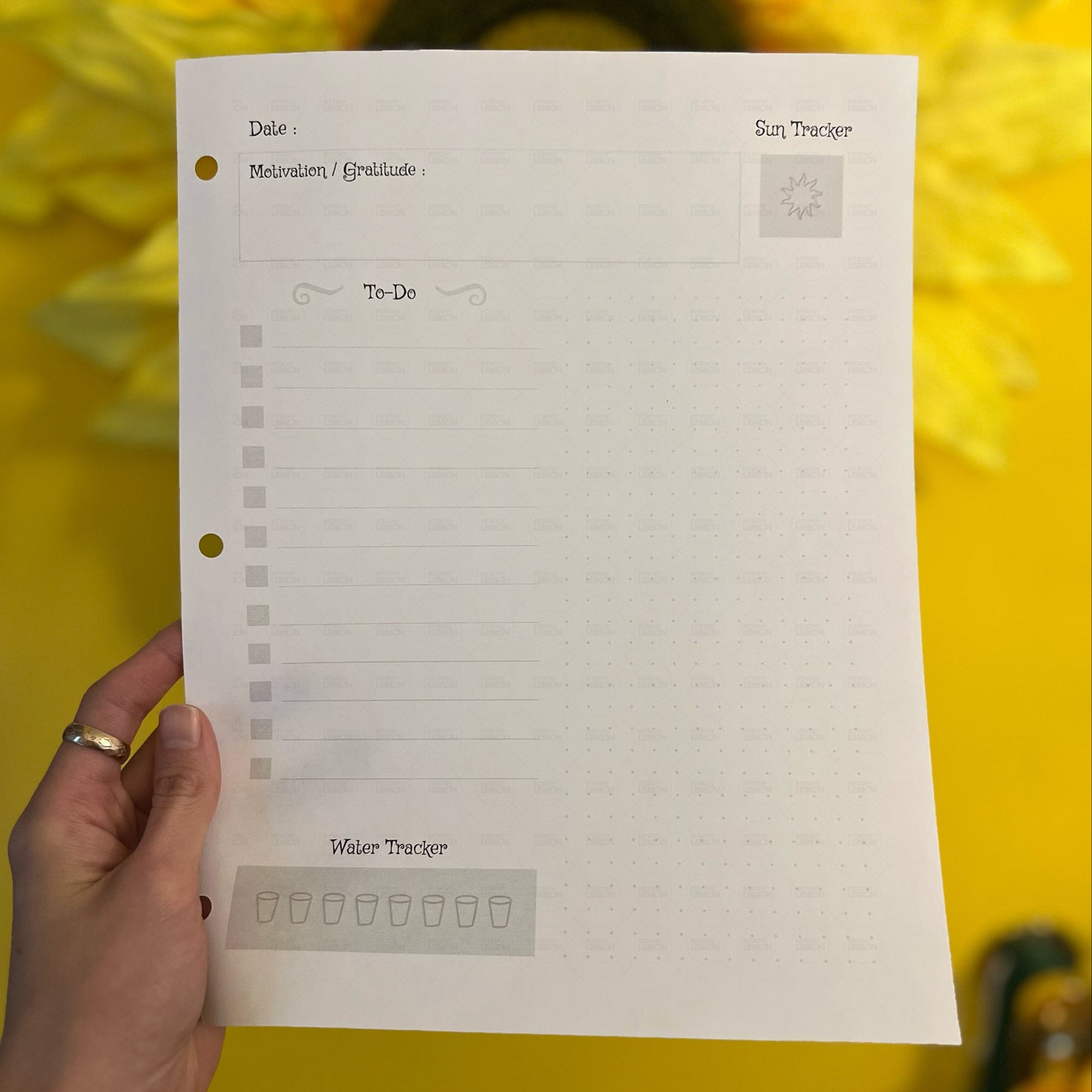 Hand holding a planner sheet with various tracking sections against a yellow background with sunflowers.